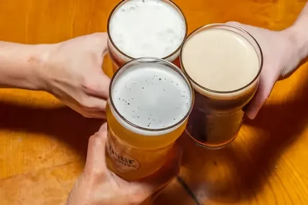 Three hands toast with different beers on a wooden table.