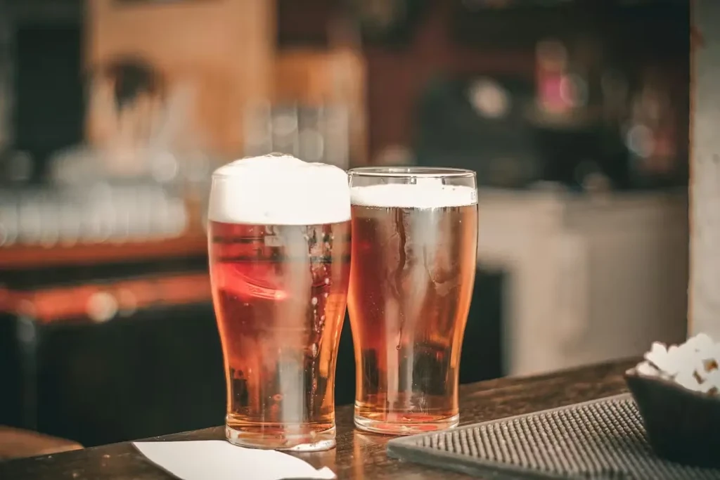 Two pints of golden beer, varying foam, on a bar with snacks.