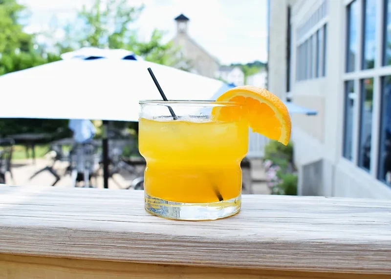 Yellow-orange cocktail with ice and orange slice on a wooden surface, outdoors.