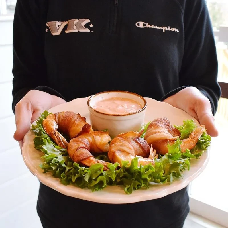 Person holds plate of bacon-wrapped shrimp, lettuce, and dipping sauce.