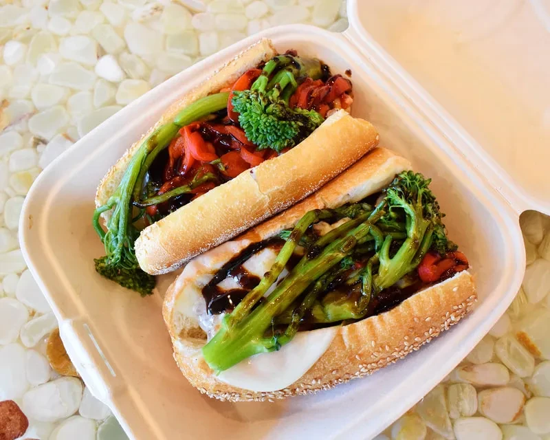 Two open-faced hoagies with vegetables and cheese in takeout container.