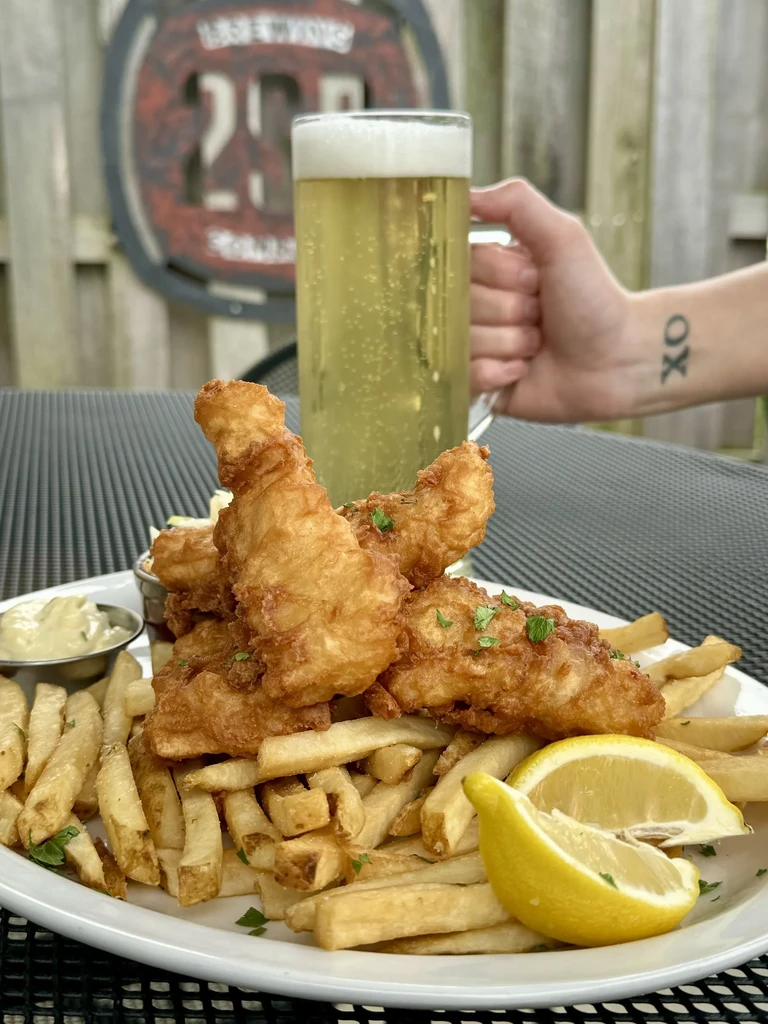 Plate of fish and chips, lemon, tartar sauce; hand holds a beer.