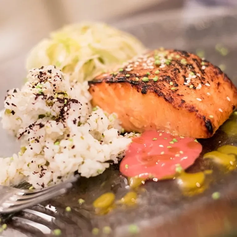 Close-up of charred salmon, white rice, green vegetables, sauces, and fork.