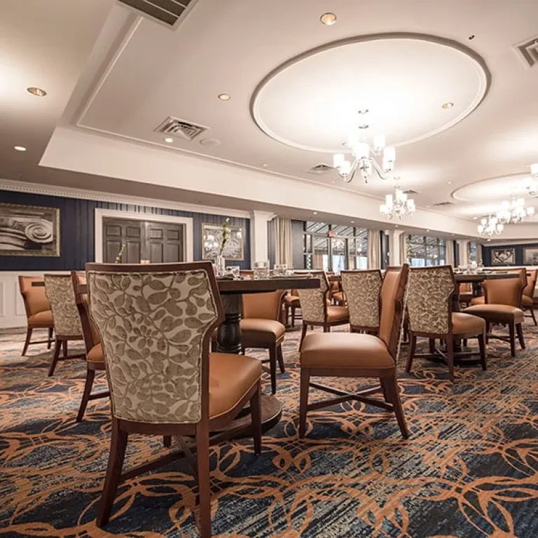 Elegant, empty dining room with dark tables, upholstered chairs, patterned carpet.