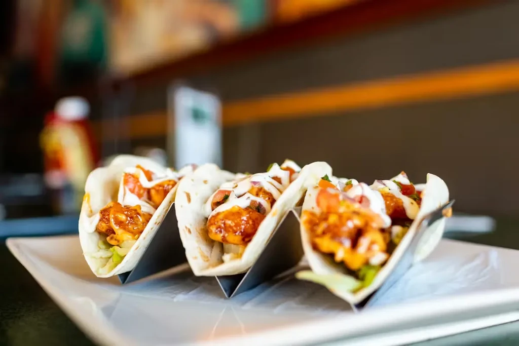 Three soft-shell tacos with orange protein, lettuce, red topping, white sauce.