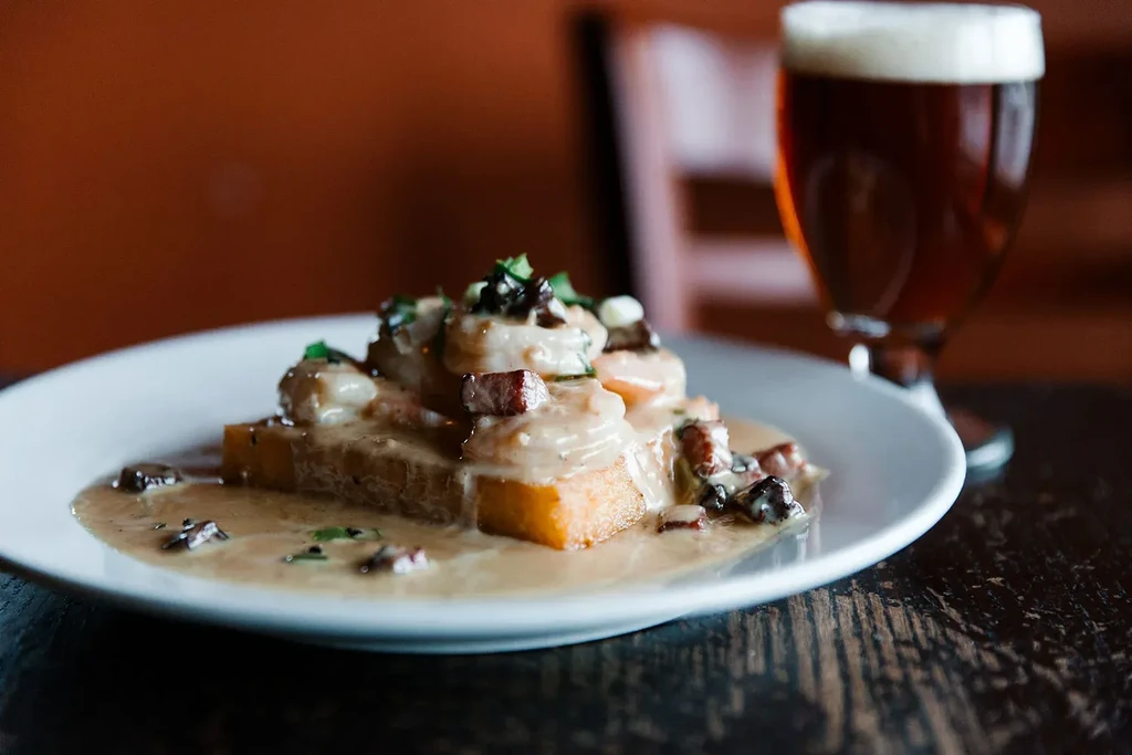 Plate of shrimp, mushrooms, and bacon over grits with a beer.