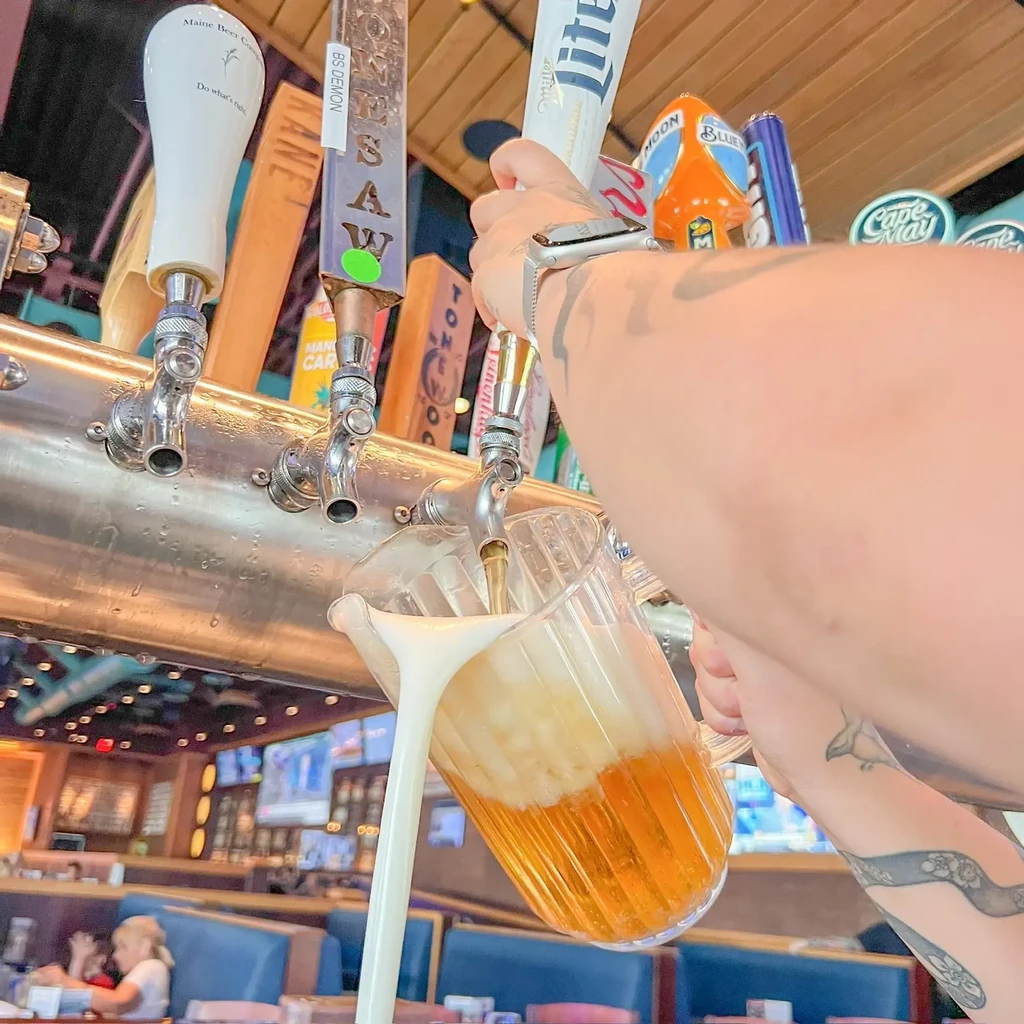 Tattooed arm pours a pitcher of golden beer from a tap.