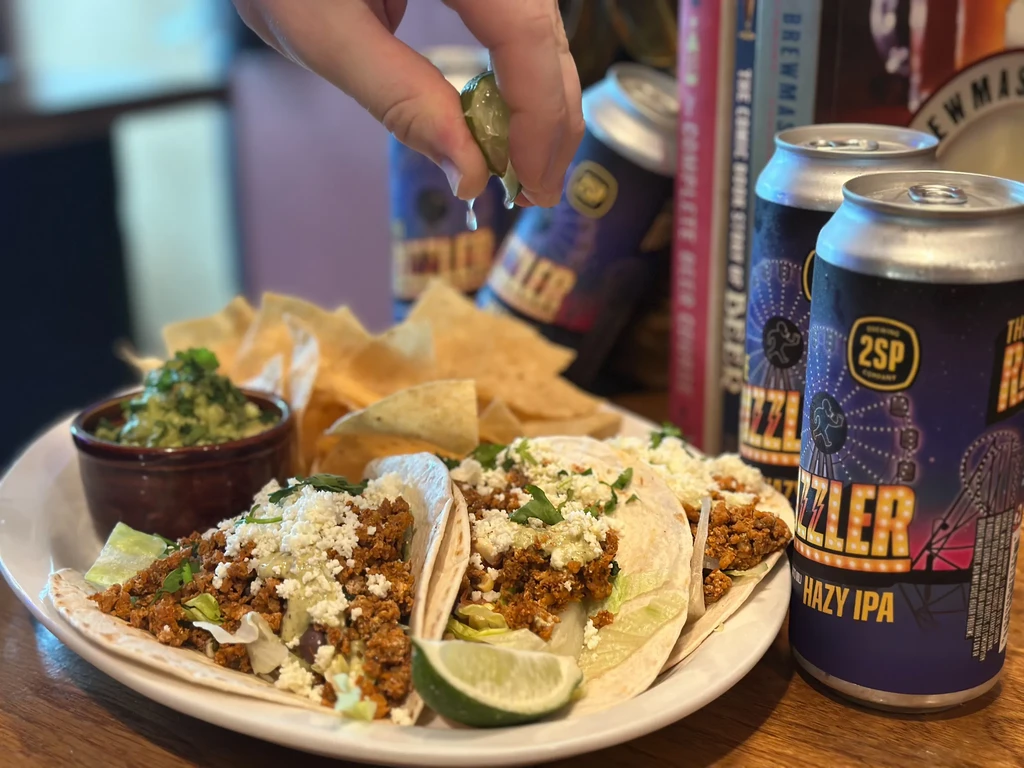 Hand squeezing lime over tacos, guacamole, chips, and IPA beer.