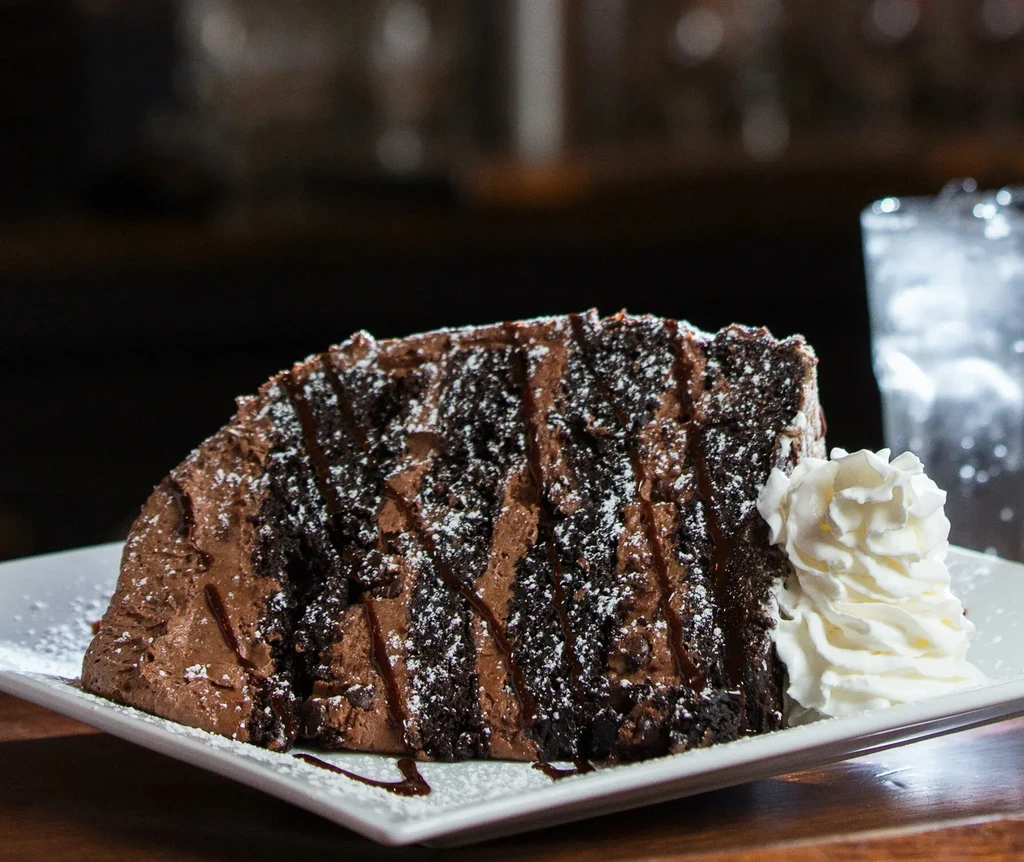 Multi-layered chocolate cake slice with frosting, sauce, powdered sugar, and whipped cream.