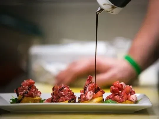 Hand drizzles dark sauce onto four red-topped appetizers.