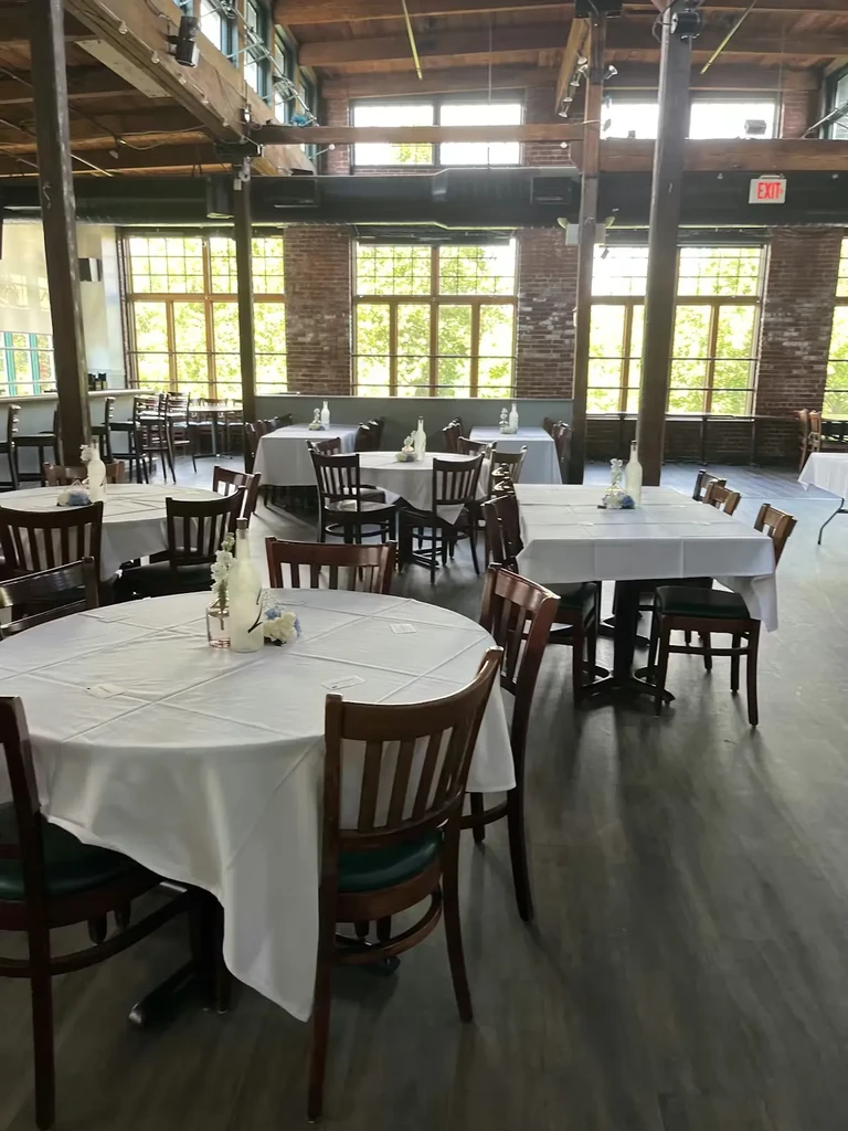 Empty restaurant hall with white tables, exposed beams, and brick walls.