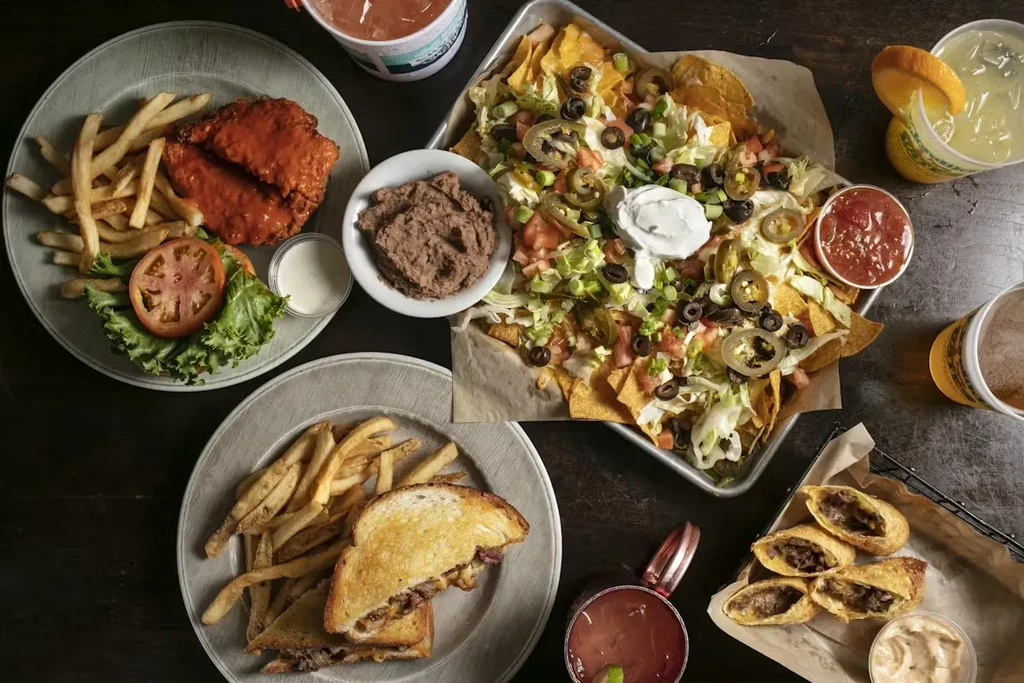 High-angle view of a dark table with nachos, sandwiches, and cocktails.