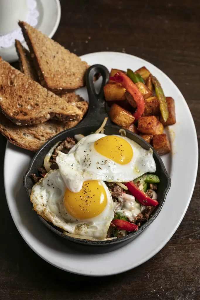 Breakfast skillet with fried eggs, hash, melted cheese, potatoes, and toast.