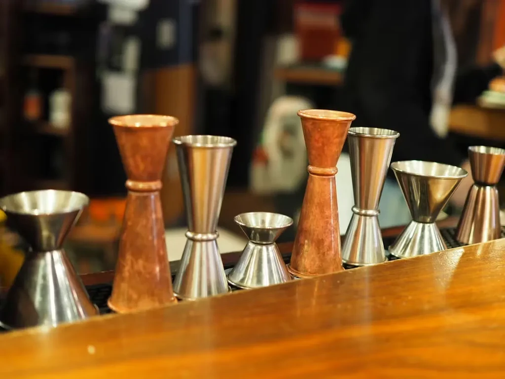 Seven copper and silver cocktail jiggers on a wooden bar.