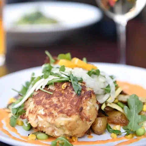 Plated crab cake garnished with greens, orange, potatoes, and corn.