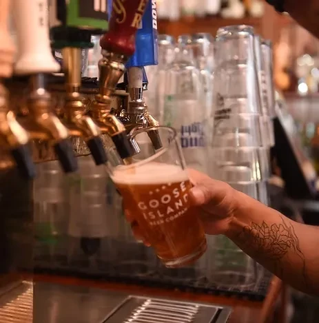 Tattooed hand holds Goose Island glass, golden beer fills from tap.