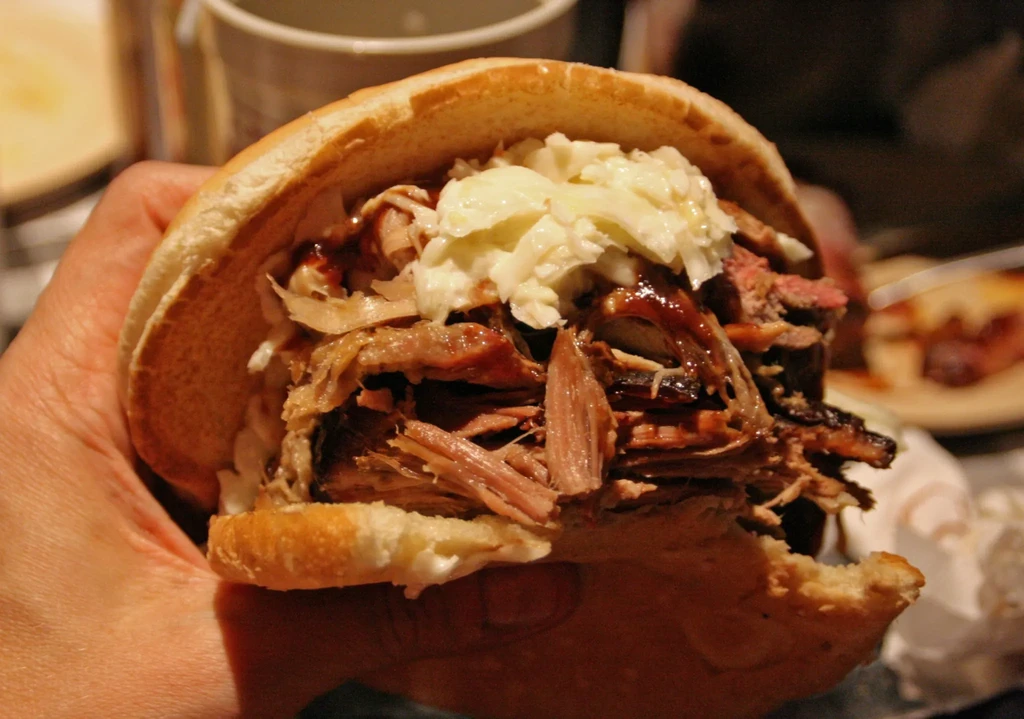 Hand holding a bitten, overflowing pulled pork sandwich with coleslaw.