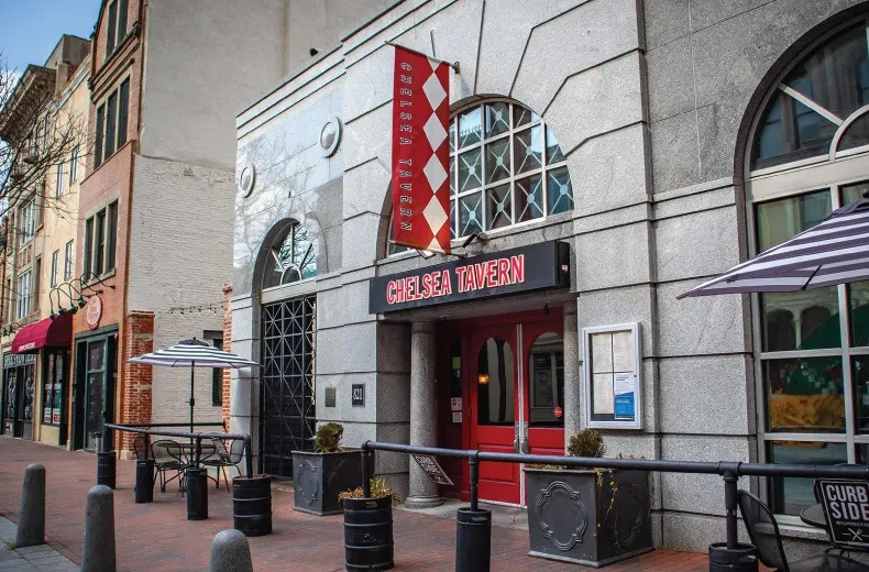 Grey stone Chelsea Tavern building with red doors and outdoor seating.
