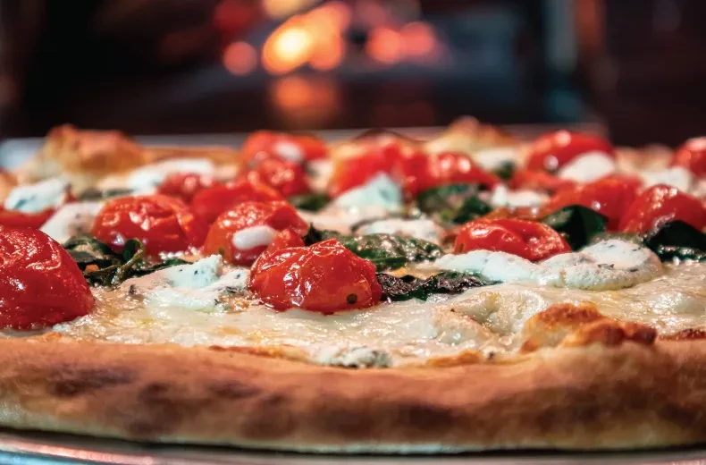 Wood-fired pizza with cheese, roasted tomatoes, and green leaves.