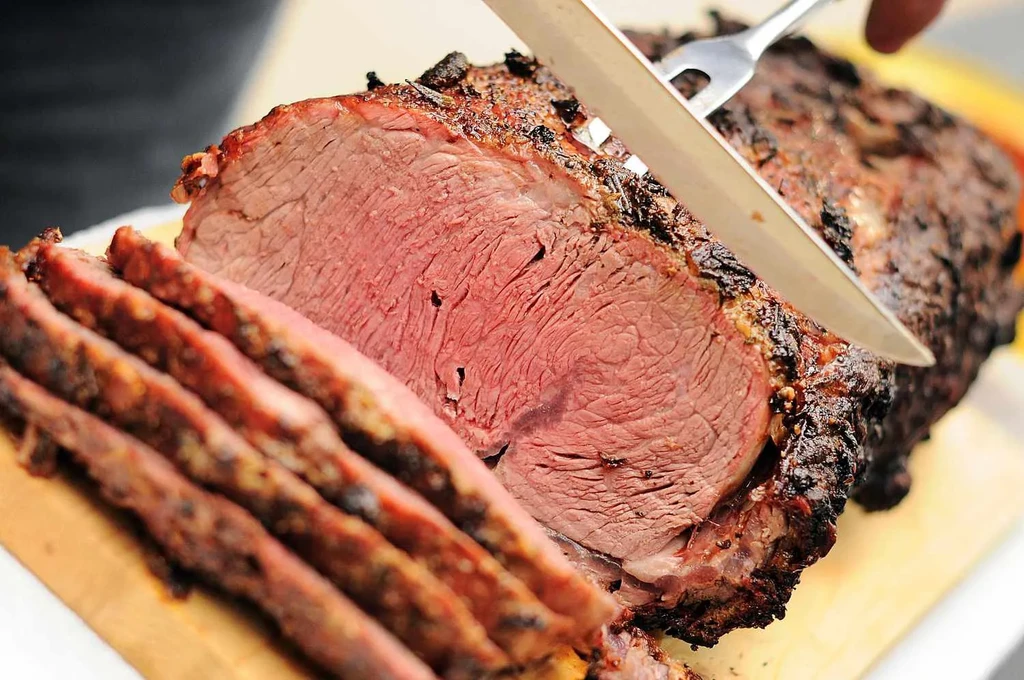 Hands carve medium-rare roast with crispy crust on cutting board.