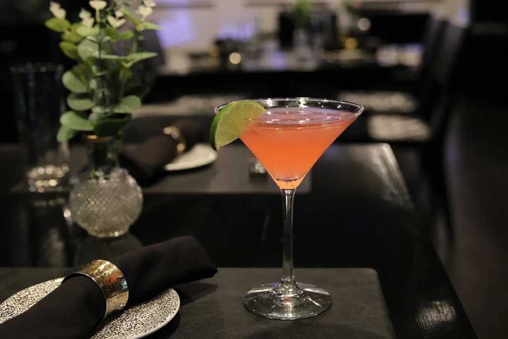 Pinkish-orange cocktail, lime, dark napkin, floral vase on a dim table.