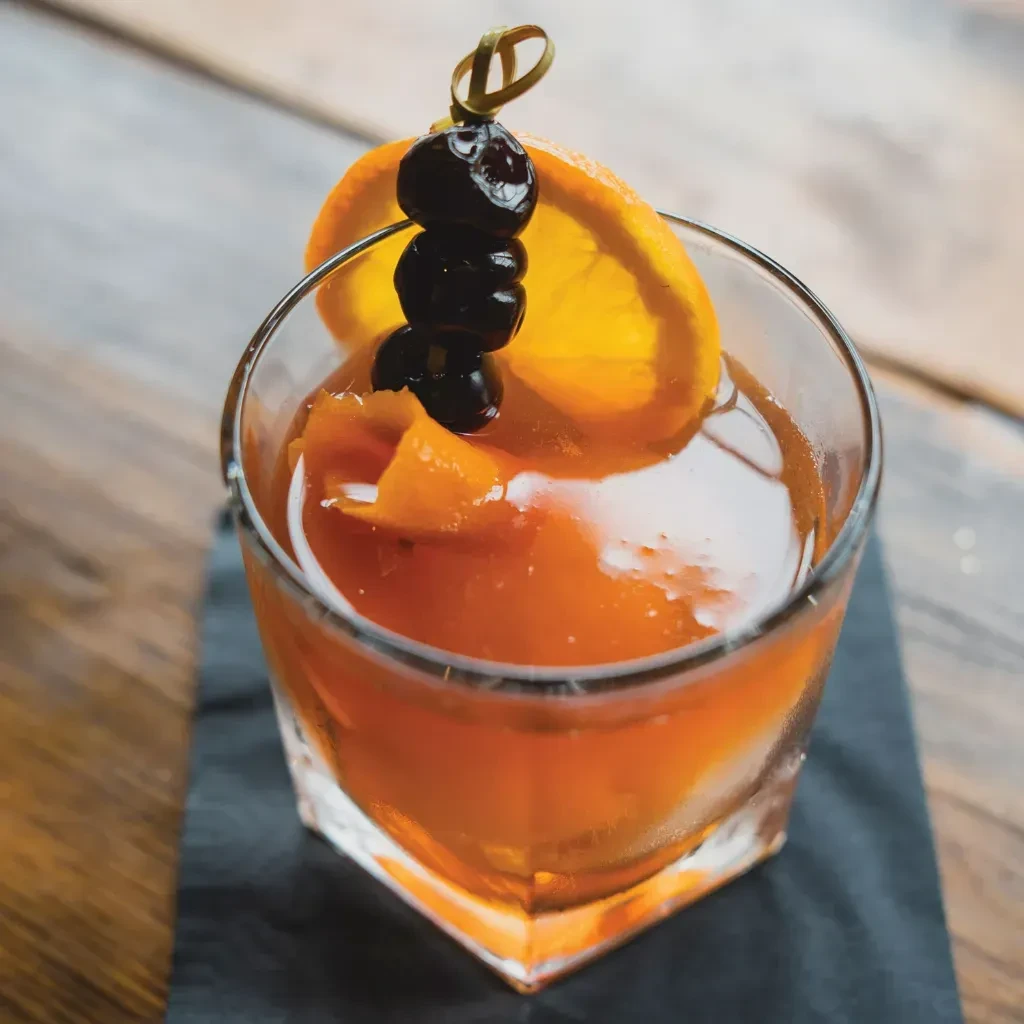 Amber cocktail in faceted glass, garnished with orange and cherries.