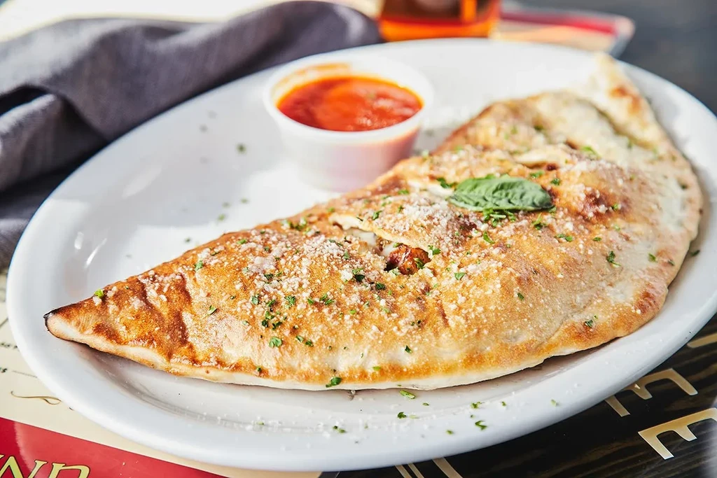 Golden-brown triangular calzone topped with cheese, herbs, and basil with marinara.
