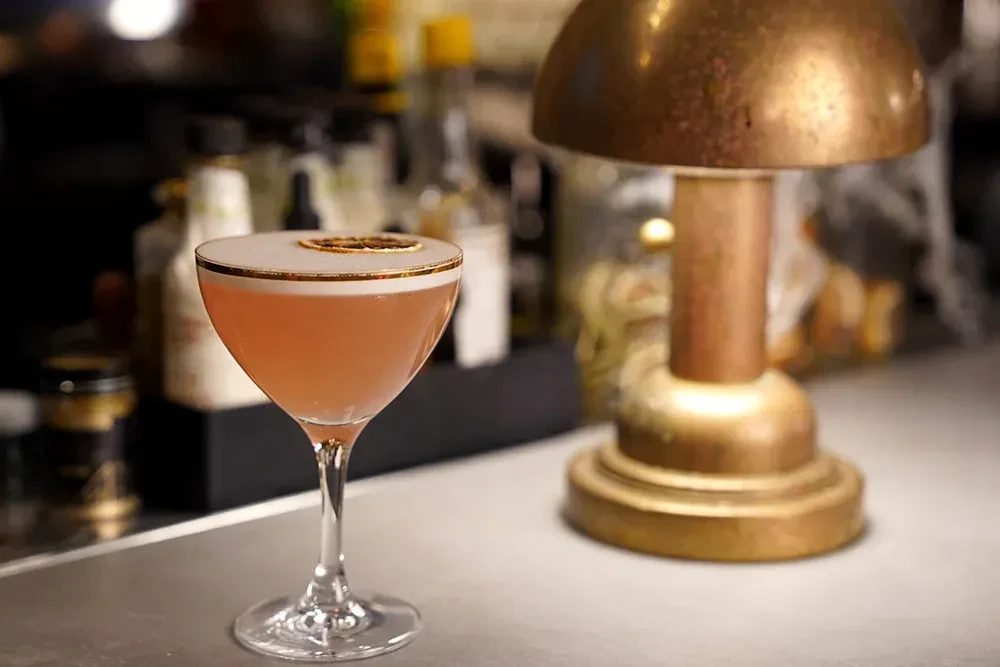 Pink cocktail with foam, dried citrus, golden rim, on bar.