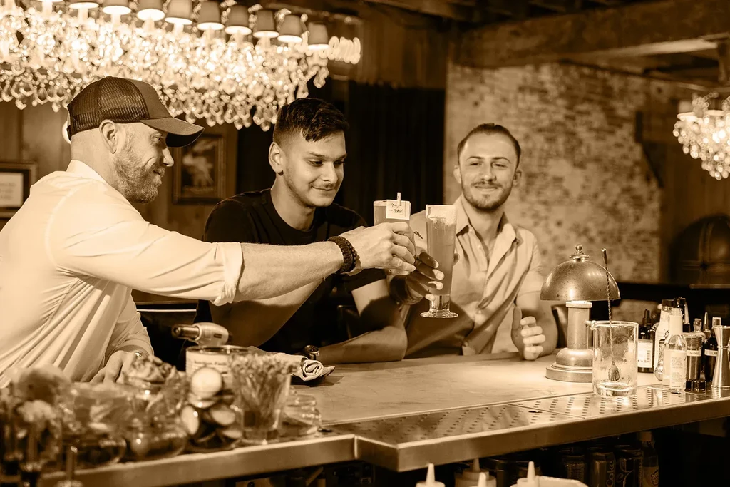 Three men smiling with drinks at a sepia-toned bar counter.