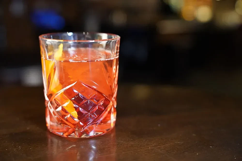 Reddish-orange cocktail in diamond-cut glass with a blurred bar background.