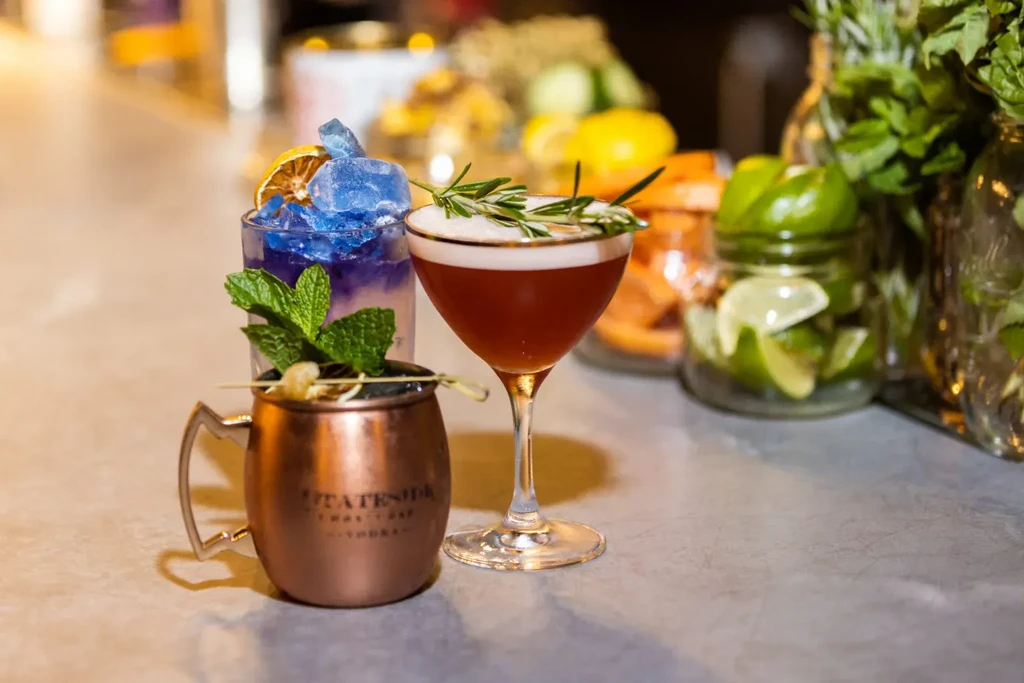 Three distinct garnished cocktails on a bar with blurred ingredients.