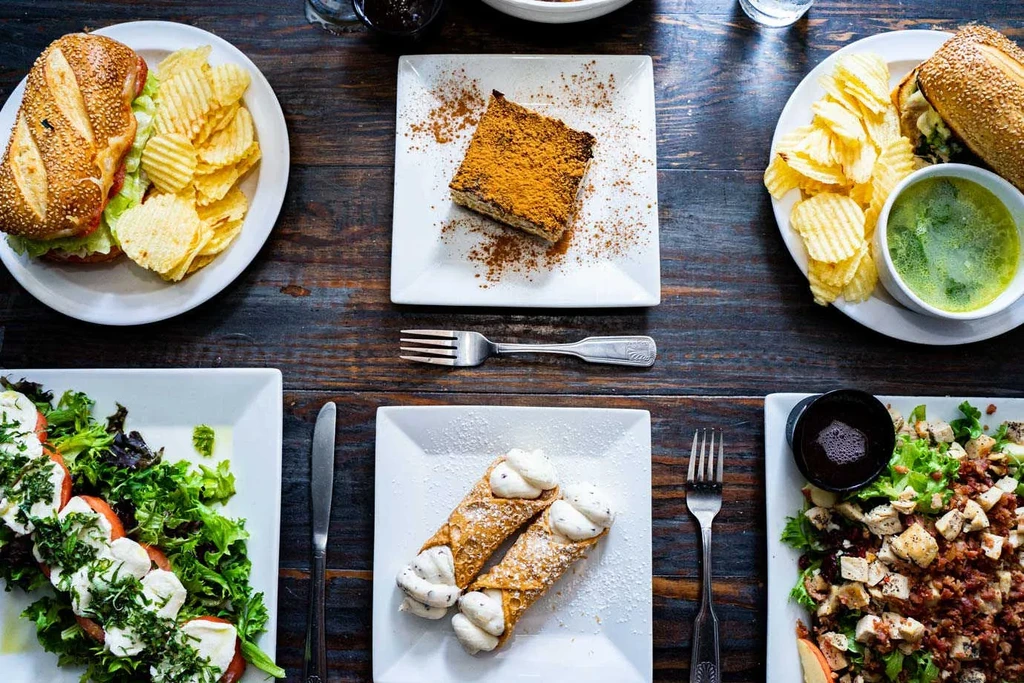 Overhead shot: six plated sandwiches, salads, desserts on dark table.