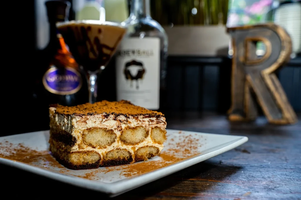 Close-up of layered tiramisu, cocoa dusted, beside a blurred cocktail.