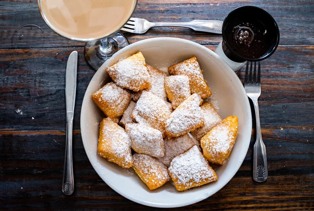 Powdered sugar beignets, drinks, and chocolate sauce on a rustic wooden table.