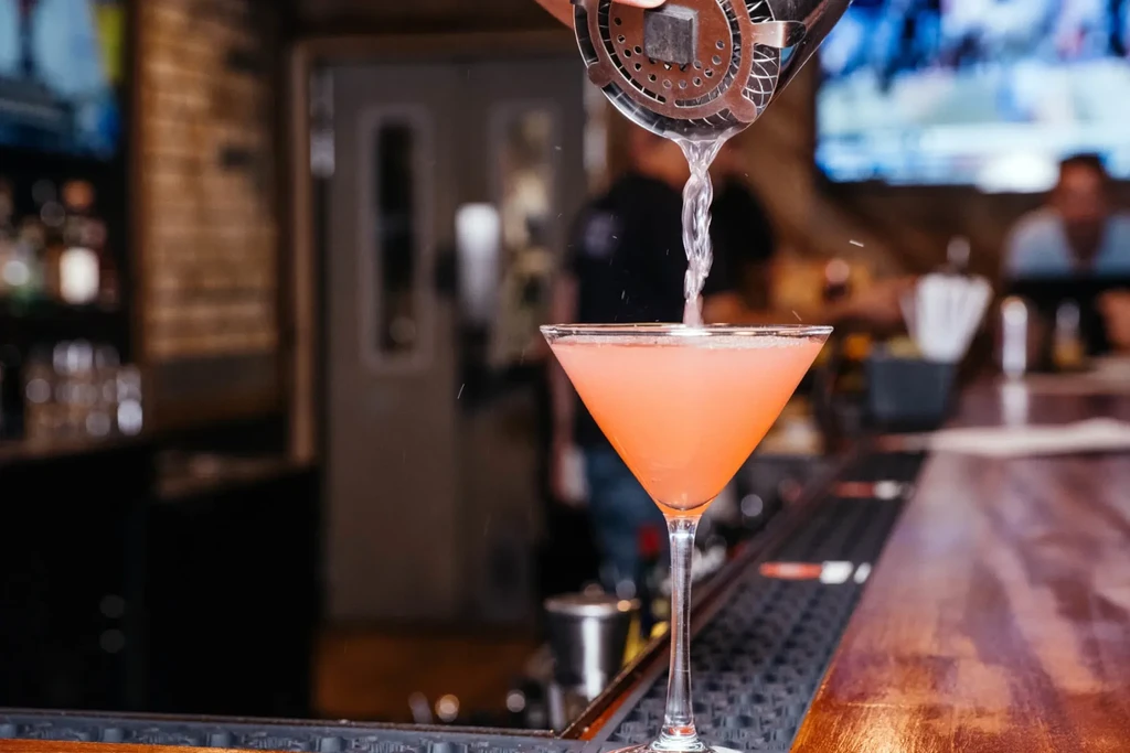 Hand pours pink cocktail from strainer into martini glass at bar.