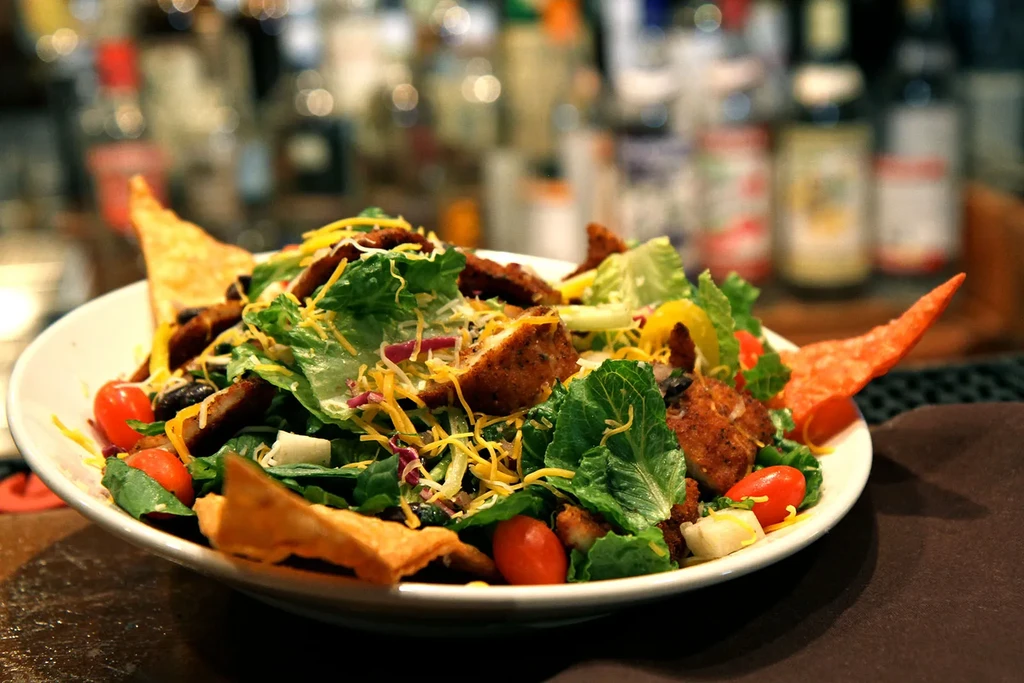 White bowl holds a salad with fried chicken, cheeses, vegetables, chips.