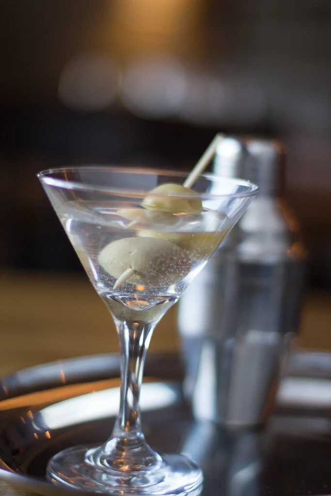 Martini glass with clear drink, two green olives, and blurred shaker.