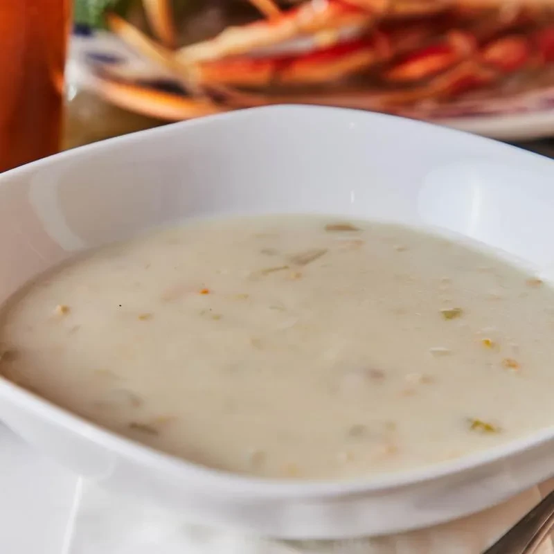Creamy chowder in a white bowl with blurred seafood and a drink.