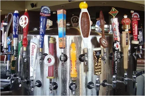 Various branded beer tap handles on a cold stainless steel bar.