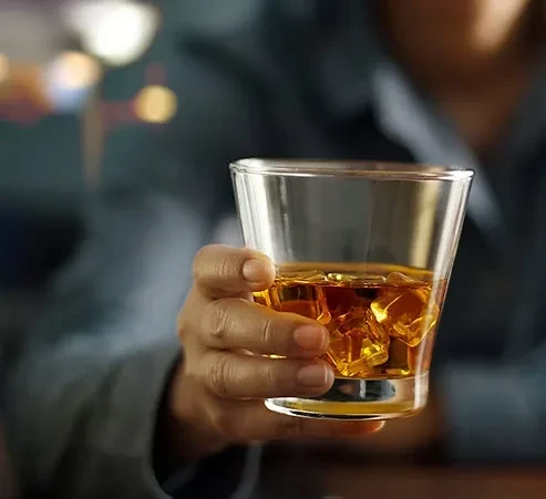Dark-skinned hand holding a clear glass of whiskey with ice.