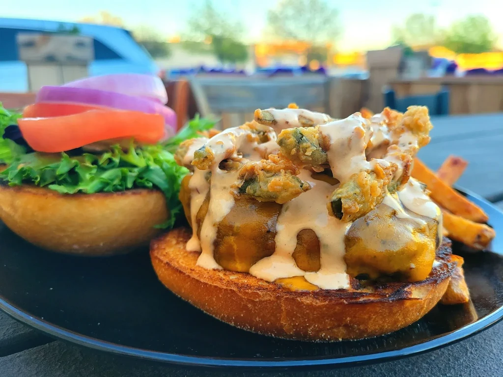 Open-faced burger topped with fried okra, cheese, lettuce, and fries outdoors.