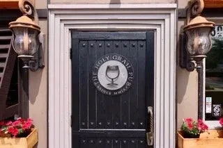 The Holy Grail Pub entrance with dark wooden door, lanterns, red flowers.