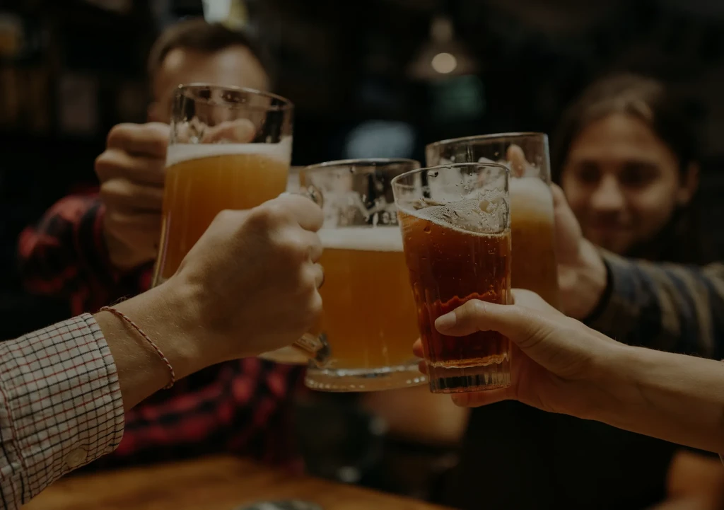 Four hands toast various golden and dark beers in a dimly lit pub.