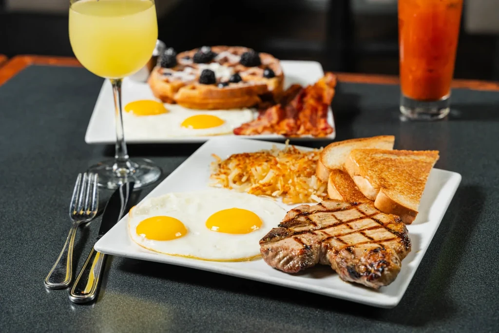 Two plates of brunch: eggs, pork chop, waffle, bacon, and drinks.