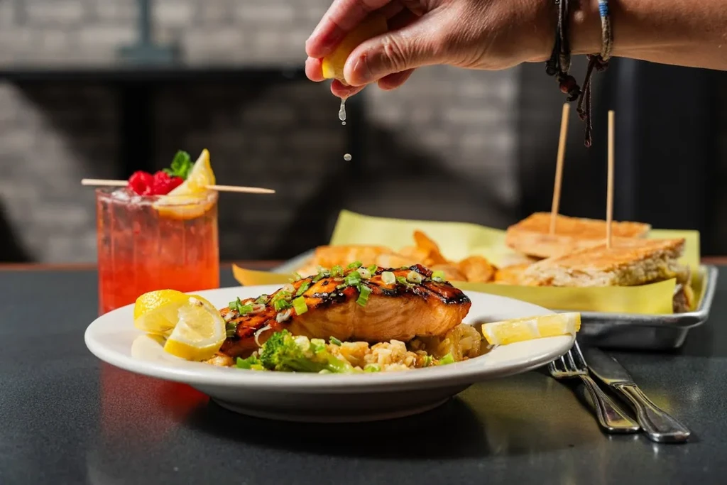 Hand squeezing lemon onto grilled salmon, broccoli, rice, and a red cocktail.