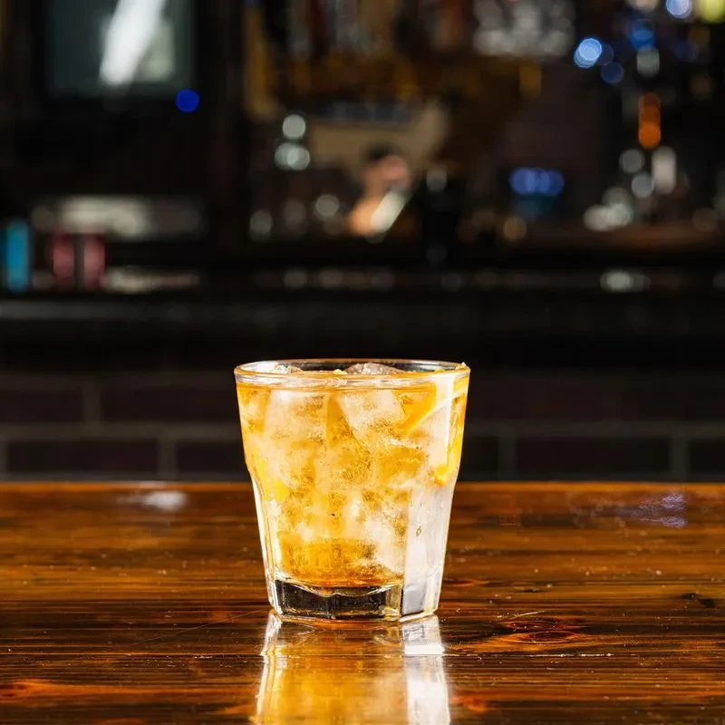 Amber drink with ice and citrus peel on a wooden bar.