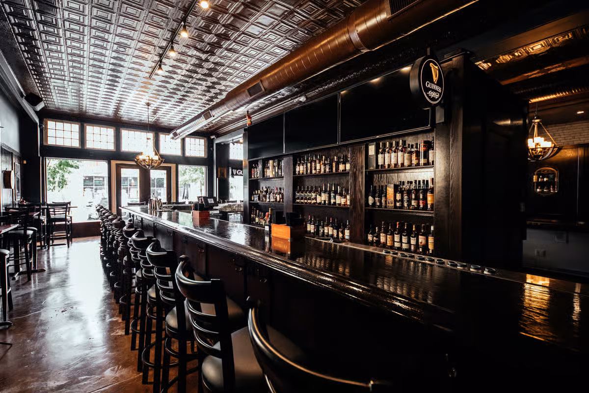 Dark pub interior with a long bar, bottles, and tin ceiling.