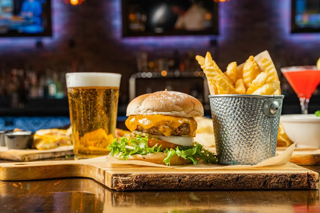 Cheeseburger, fries, beer, and red cocktail on a wooden bar counter.