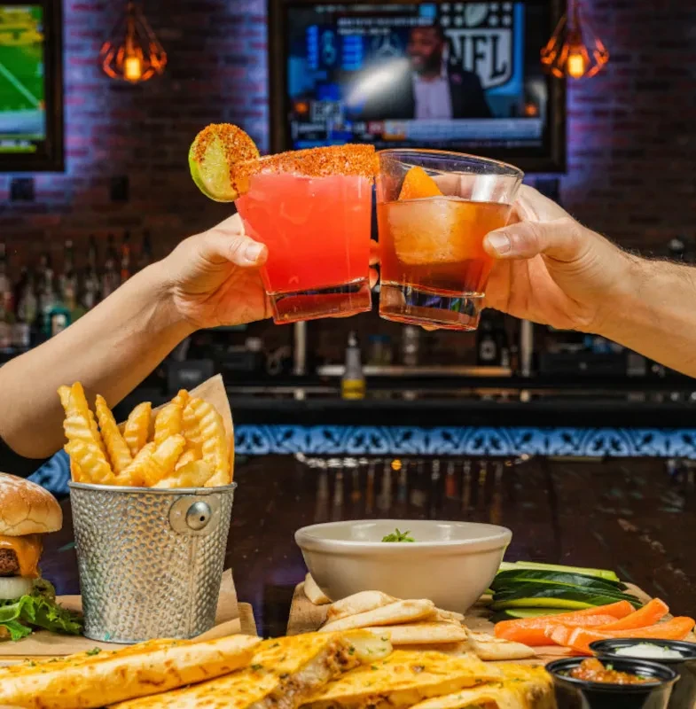 Two hands clinking cocktails, abundant bar food, and NFL on TVs.