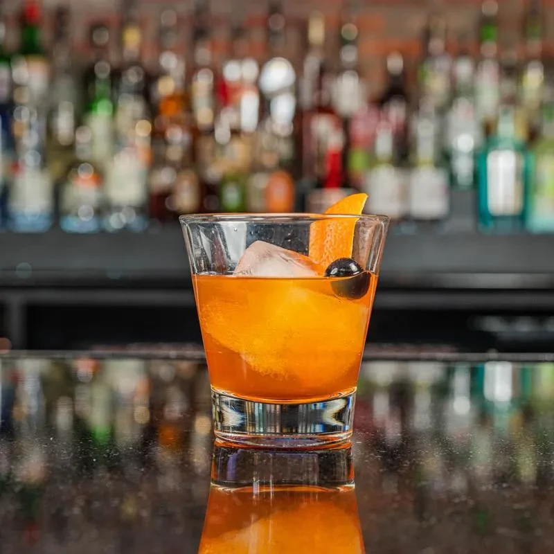 Amber cocktail with ice, orange peel, and cherry on a bar.